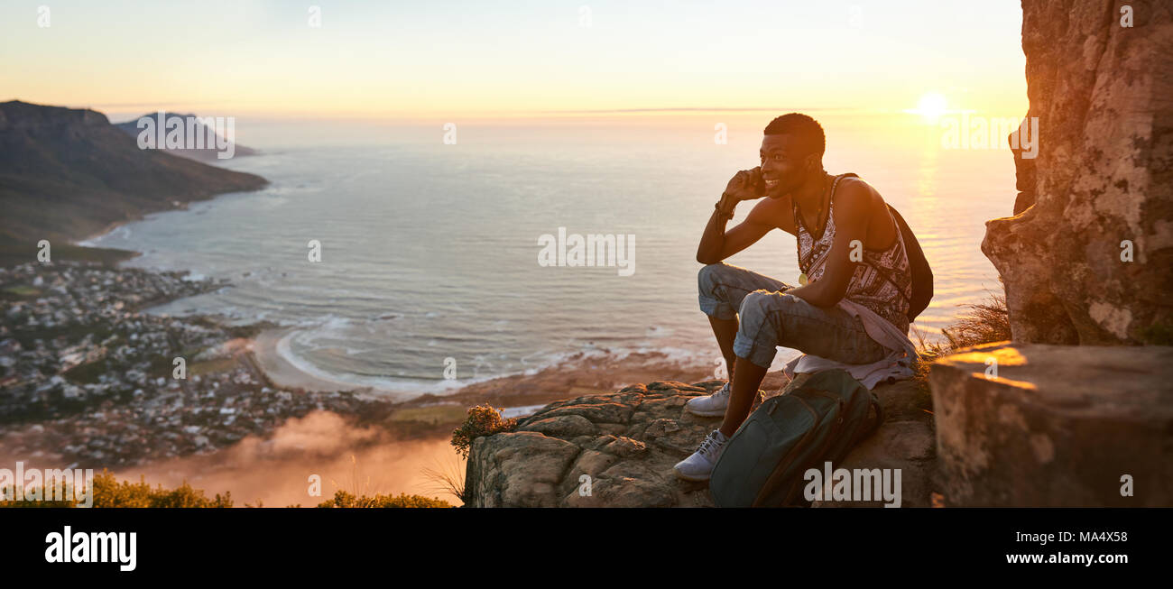 Panoramic image of a young black man sitting on the top of a mountain talking on a mobile phone during an amazing sunset, with a beautiful view of the Stock Photo