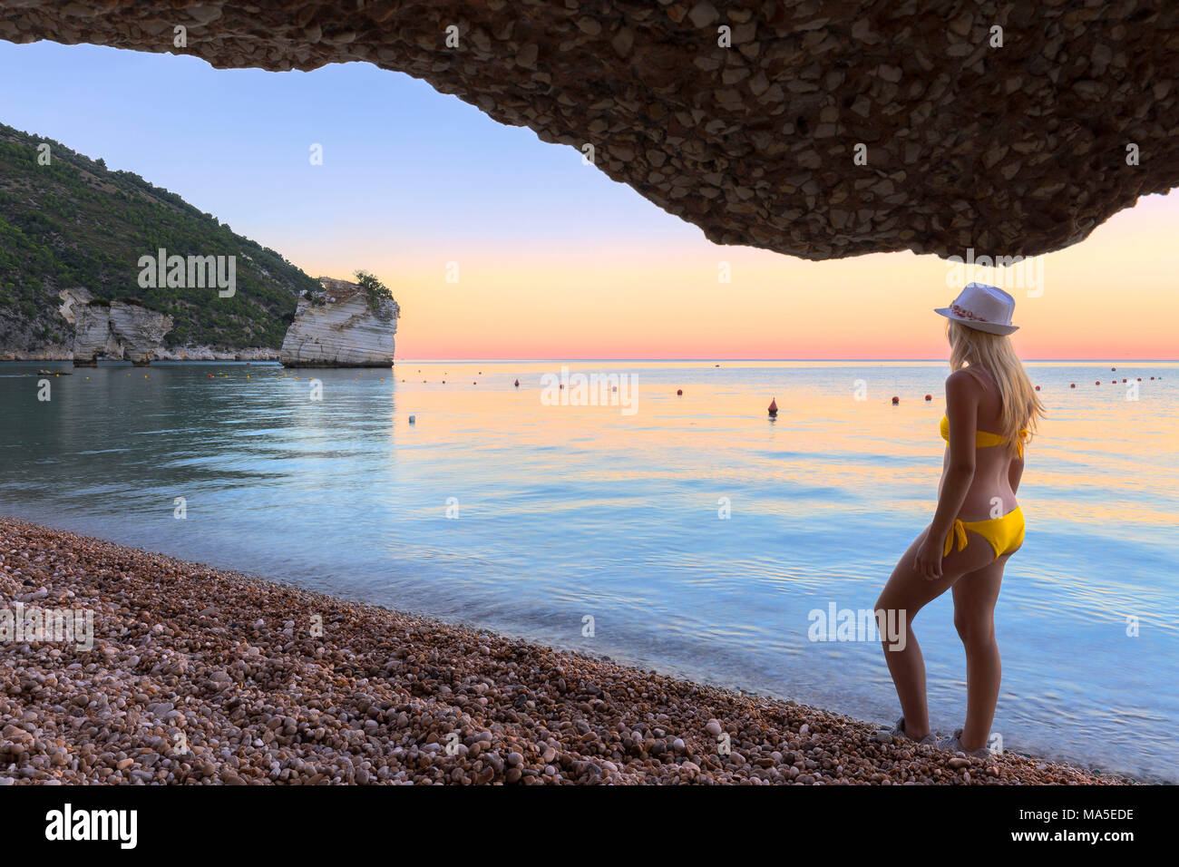 Young girl in swimsuit looks at the sunset on Faraglioni Beach(Baia delle Zagare), Mattinata, Apulia(Puglia), Italy. Stock Photo