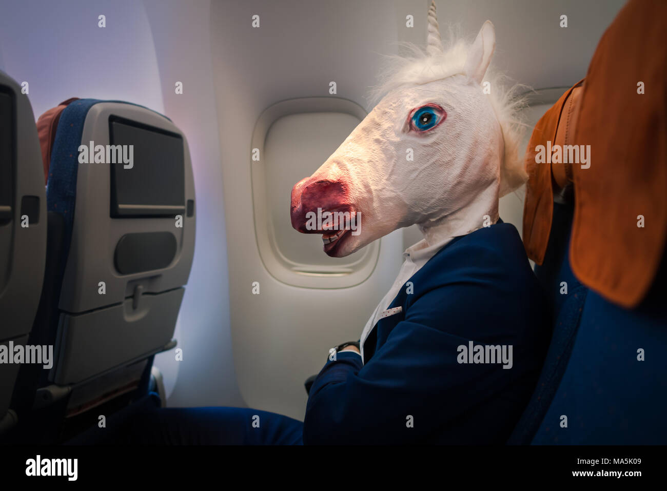 Strange young man in funny mask travels by plane. Unusual passenger in elegant suit sits alone inside the aircraft and ready to flight away. Stock Photo