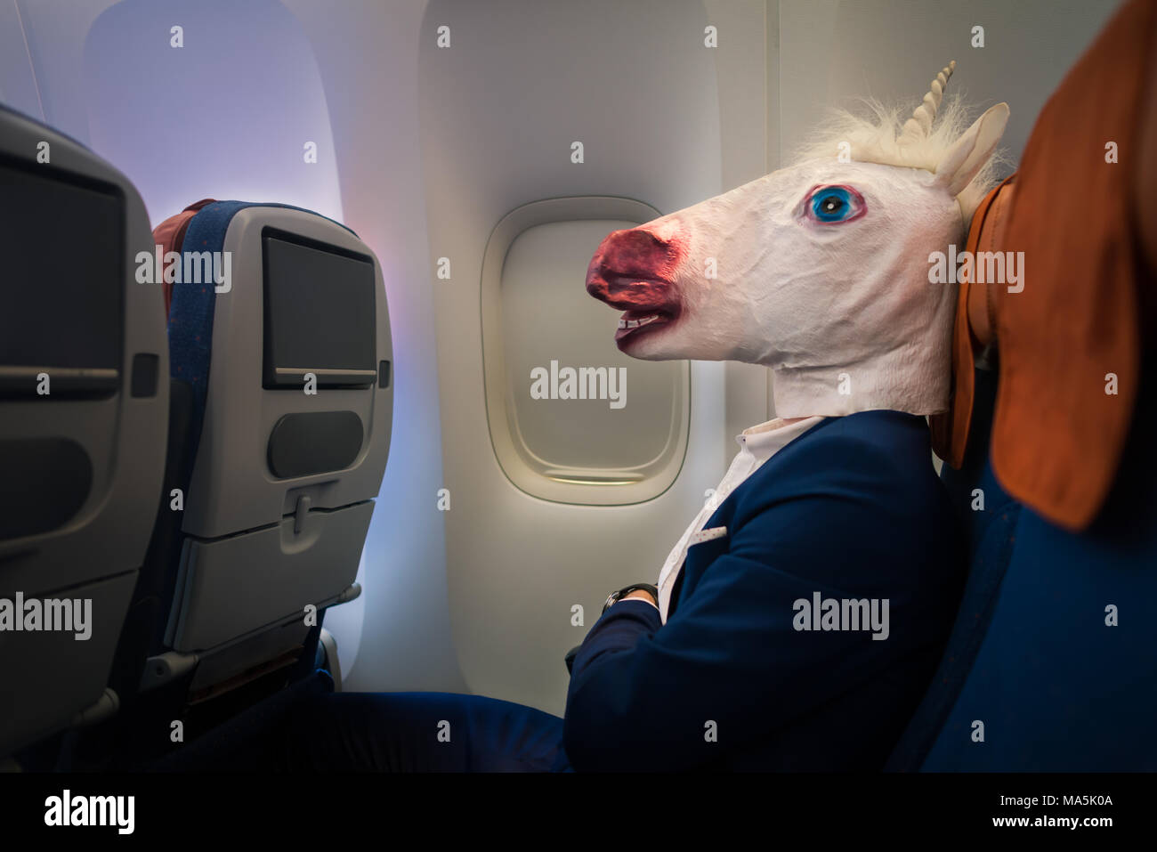 Unusual passenger in elegant suit sits alone inside the aircraft and ready to departure. Young man in funny mask travels by plane Stock Photo