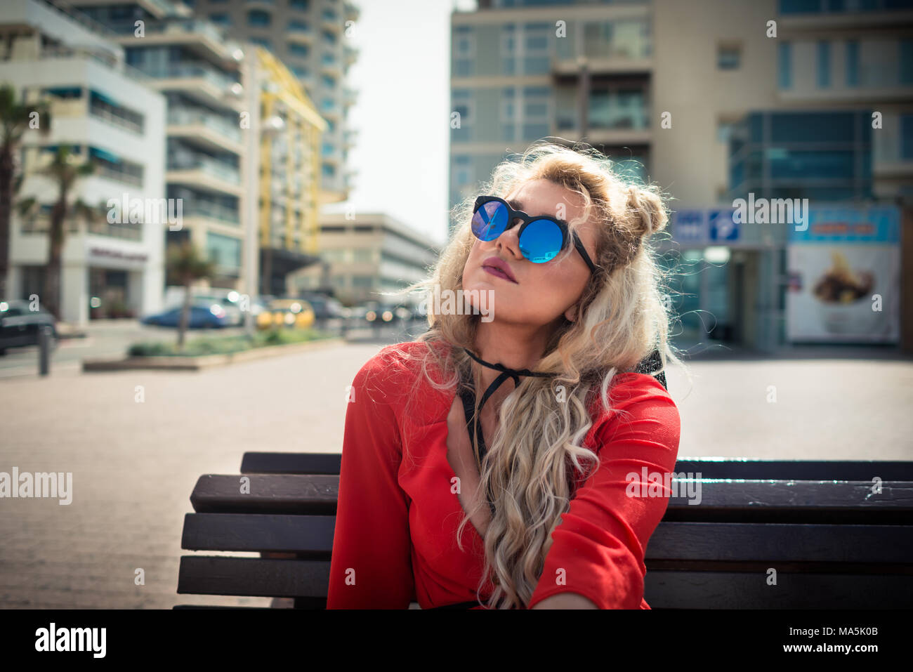 Young serious woman in red dress and sunglass sits on on the bench on background of cityscape. Pensive blonde girl is looking to sky in sunny day Stock Photo