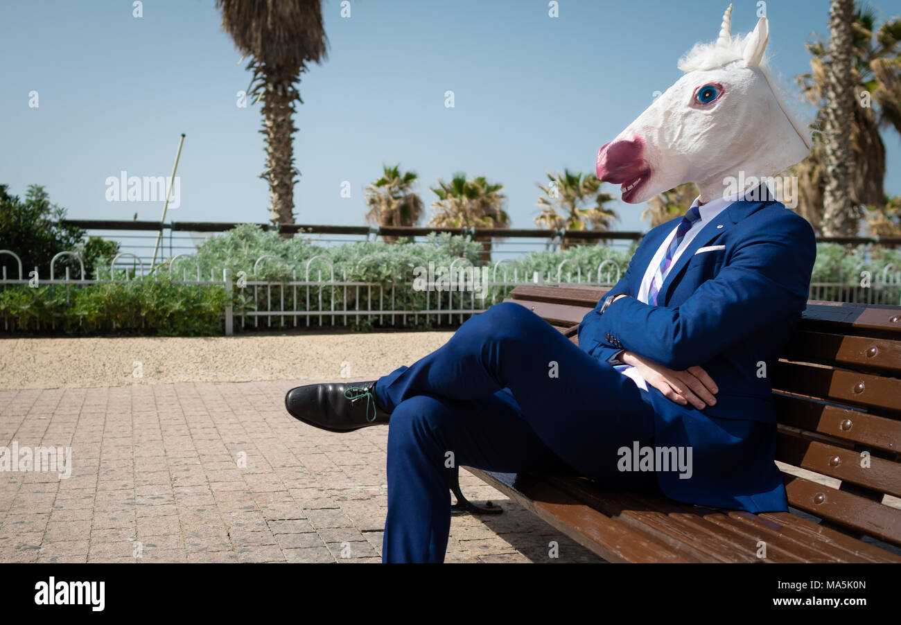 Unusual manager in elegant suit sits on the bench on city street. Young strange man in funny mask have a break in park. Unicorn is enjoying outdoors Stock Photo