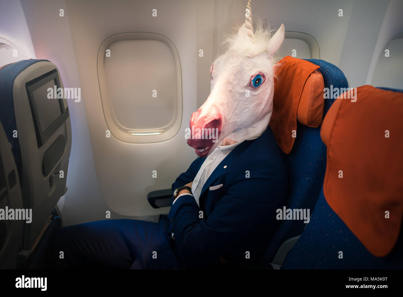 Young funny man in mask travels by plane. Unusual passenger in suit sits alone in aircraft and ready to take off. Freaky traveler unicorn enjoy flying Stock Photo