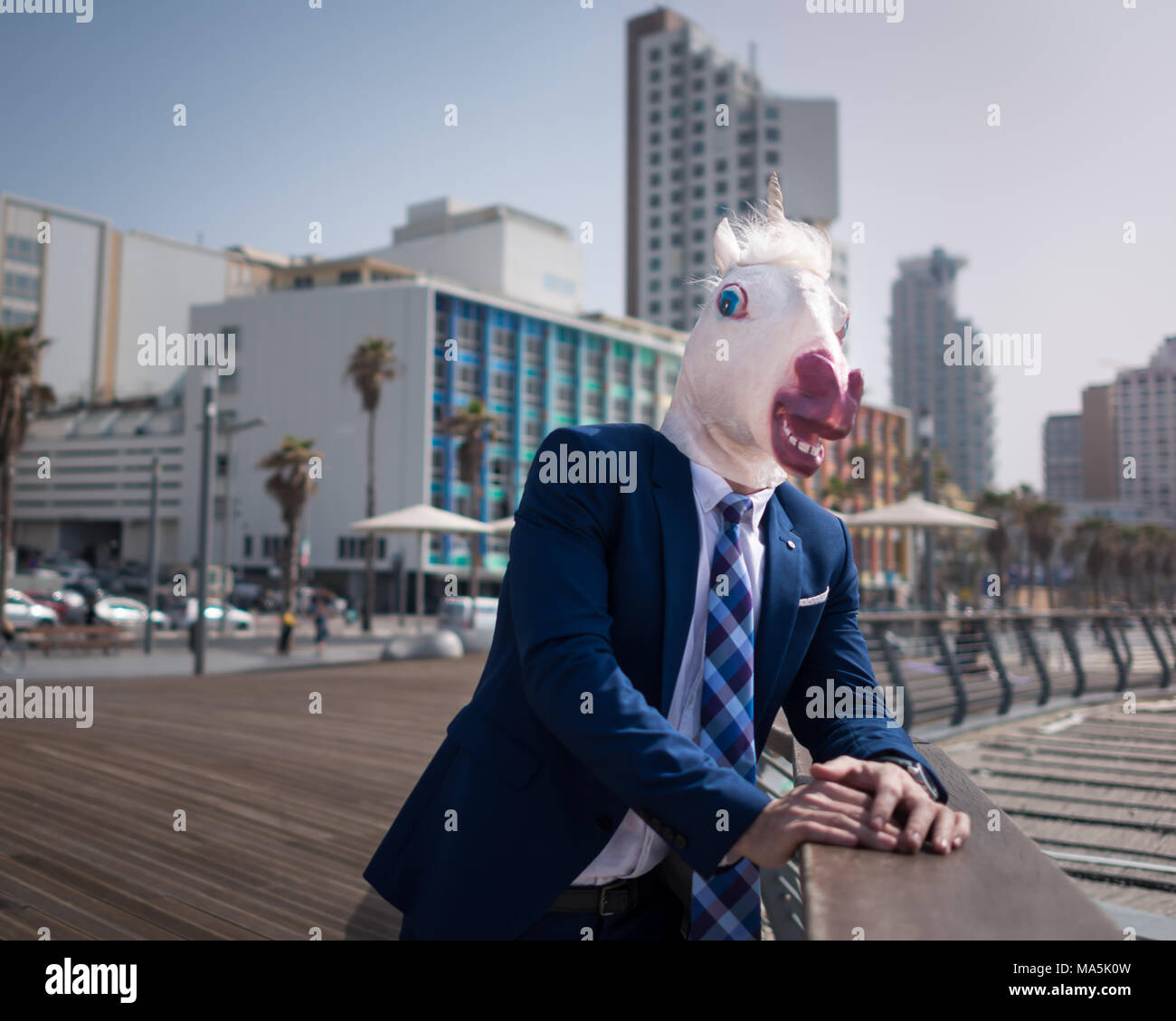 Unusual young man in suit stands on city waterfront. Strange guy in mask relaxes on city promenade. Unicorn enjoys vacation on background of cityscape Stock Photo
