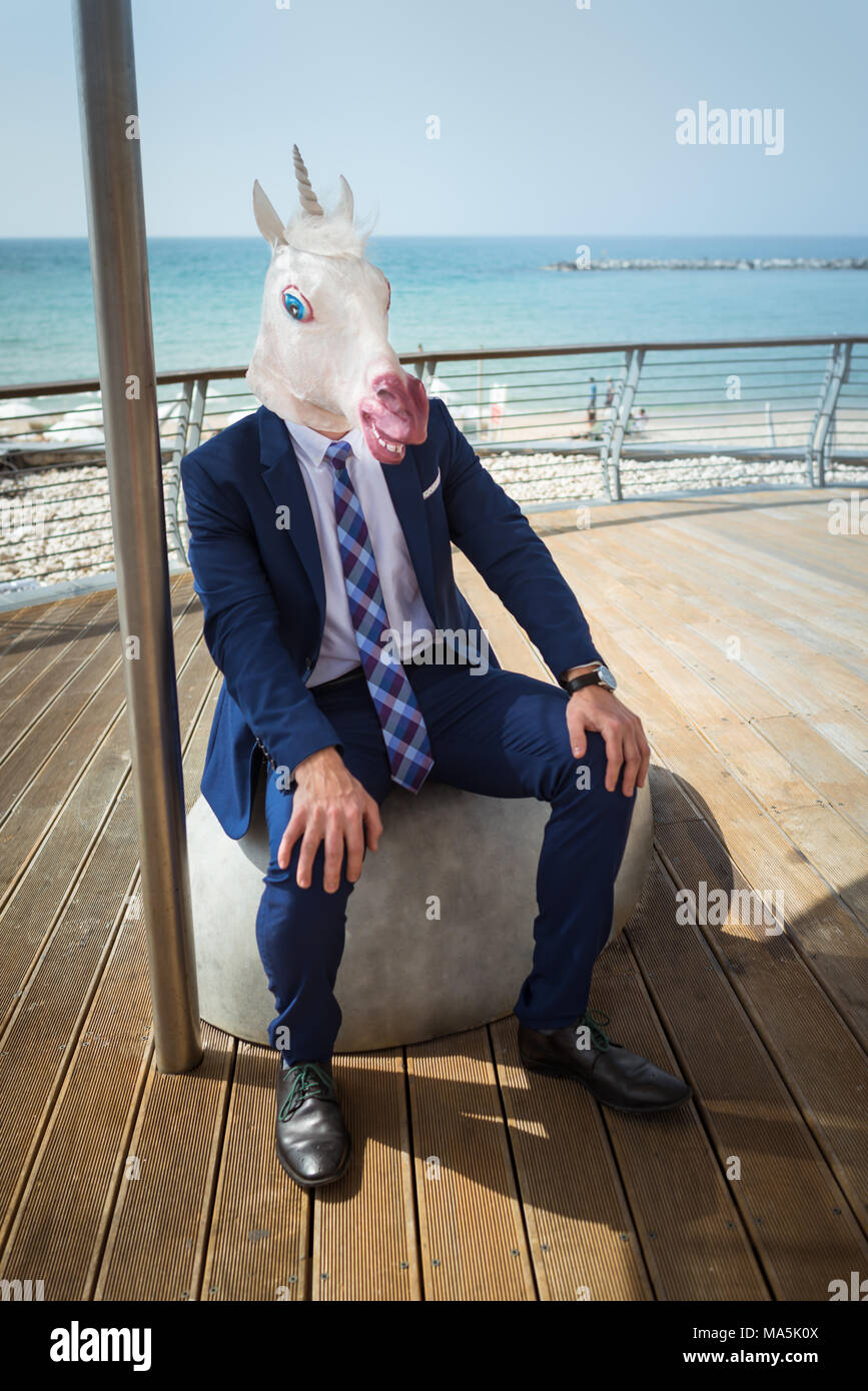 Strange guy in elegant suit sits on the city waterfront. Young unusual man in funny mask is resting on city promenade. Unicorn on background of sea Stock Photo