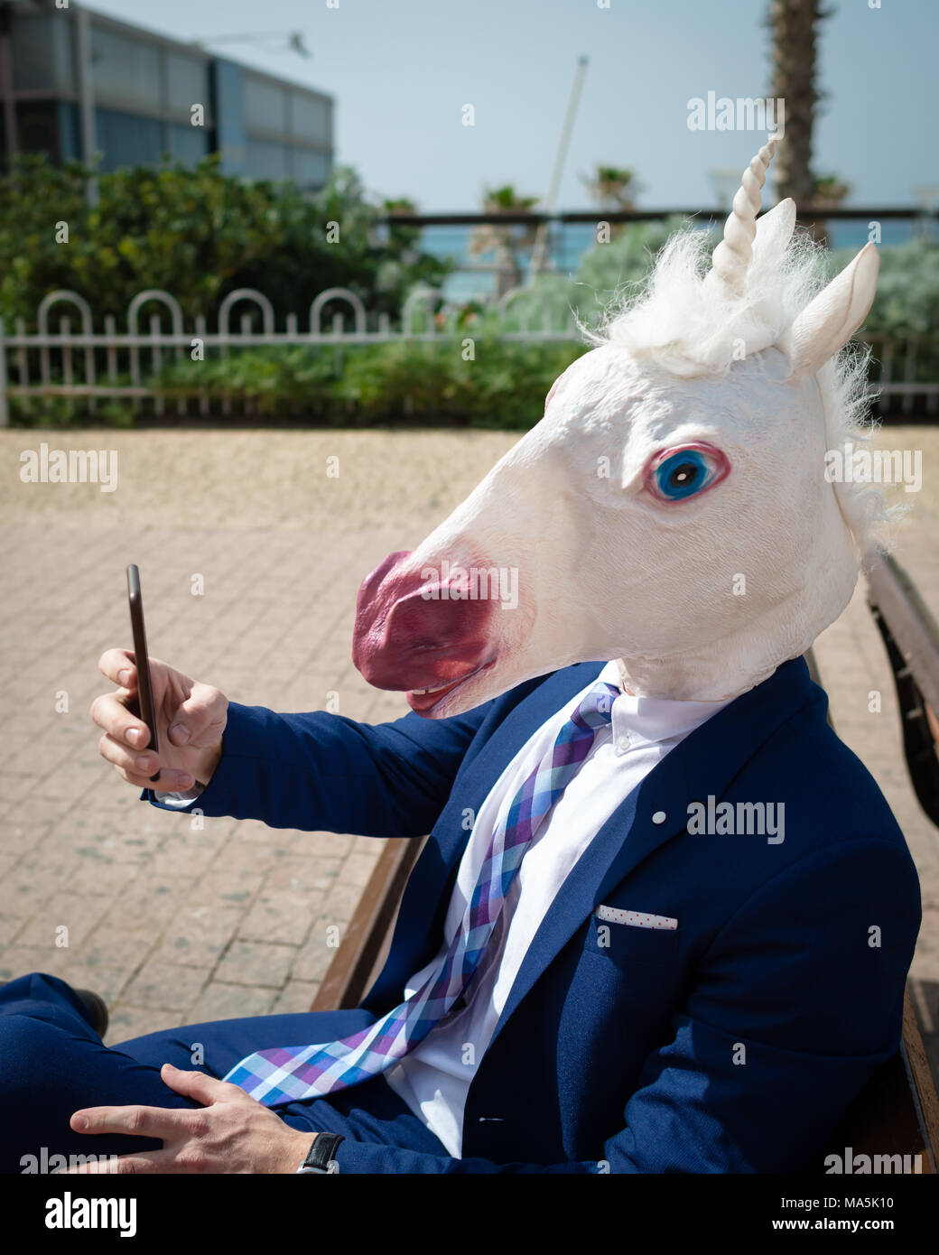 Unicorn makes a photo by phone. Young unusual man in funny mask and elegant suit is enjoying warm summer day. Strange guy sits on bench Stock Photo