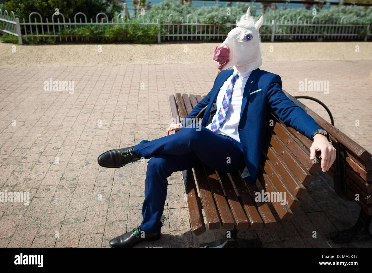 Young elegant man in funny mask and suit have a break in park. Unusual guy in sits on the bench on the city street. Unicorn is enjoying outdoors Stock Photo