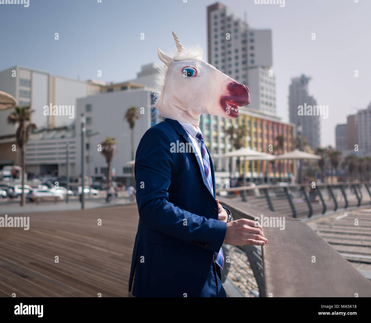 Unusual manager in funny mask and suit stands on city promenade. Young stylish man on the background of cityscape. Unicorn relaxes in warm sunny day Stock Photo