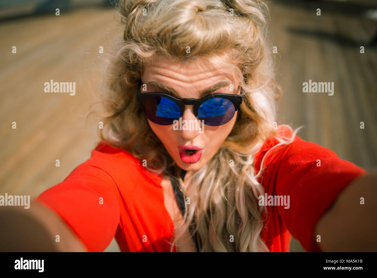 Close-up view of young woman in red dress with surprise on her face at the city waterfront. Elegant blonde girl take a selfie on wooden promenade Stock Photo