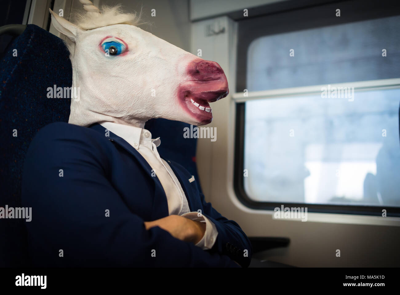 Strange passenger in suit sits alone inside the train. Young unusual man in funny mask in journey. Unicorn love to travel and adventures Stock Photo