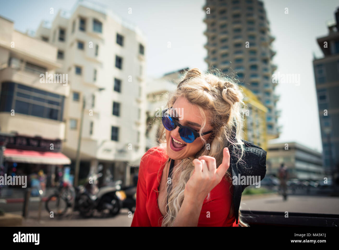 Young cute woman in red dress and sunglass sits on on bench on background of cityscape. Trendy blonde girl laughing and active gesticulate Stock Photo