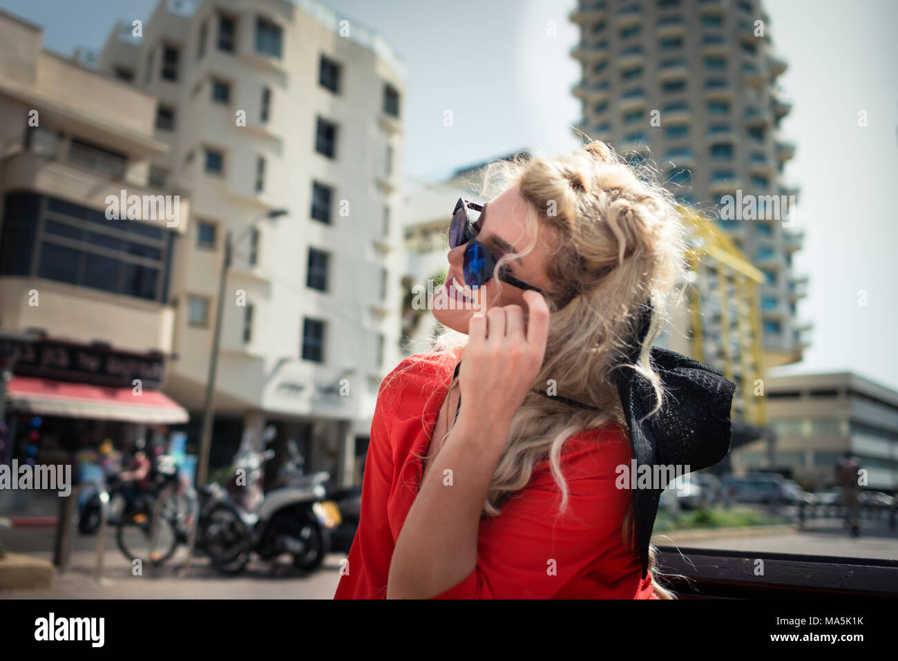Young woman in red dress and sunglass sits on bench on background of cityscape. Elegant blonde girl with smile on her face is enjoying warm summer day Stock Photo