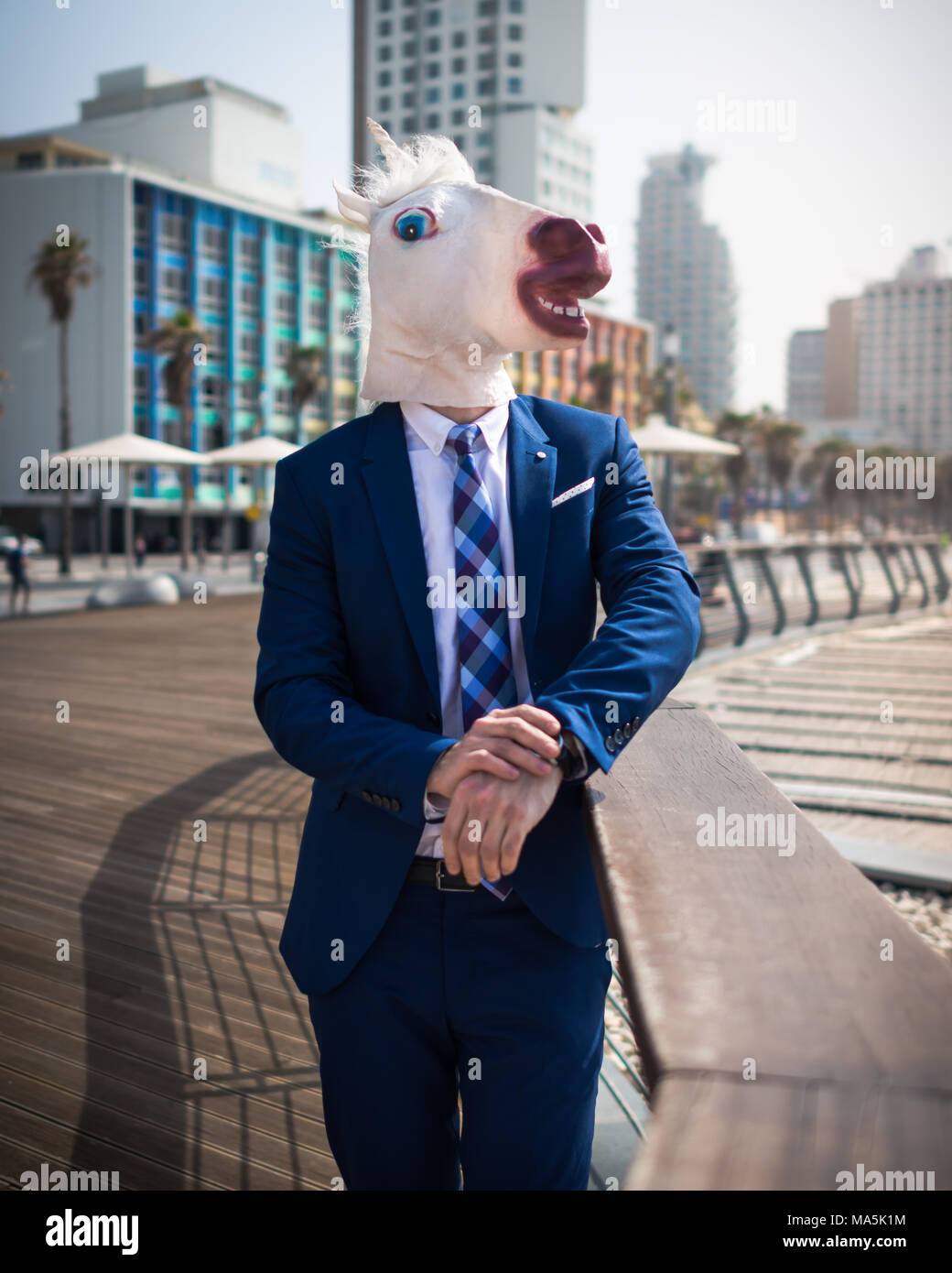 Unusual young man in suit stands on the city waterfront. Strange guy in funny mask relaxes on city promenade. Unicorn on background of cityscape Stock Photo