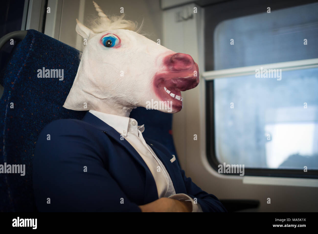 Unusual passenger in elegant suit sits alone inside the train and ready to travel. Young serious man in comical mask. Funny unicorn traveler Stock Photo