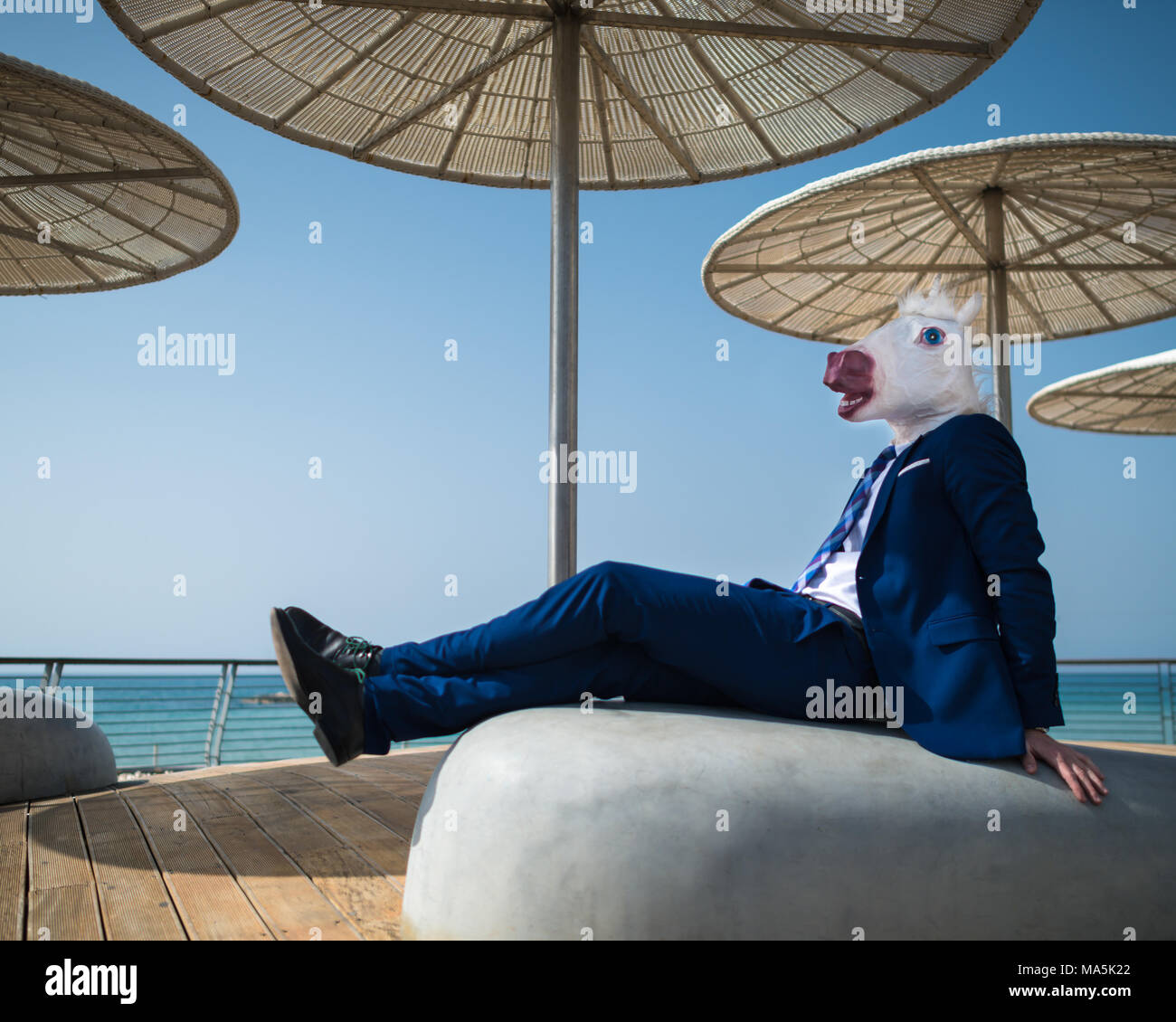 Young man in suit sits under umbrellas on city waterfront. Unusual manager in funny mask have a break. Unicorn is enjoying warm weather Stock Photo