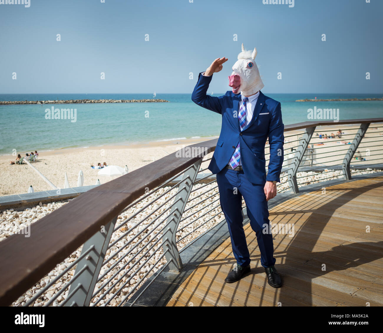 Young man in mask and suit salutes to somebody. Unicorn stands on city promenade. Unusual manager relaxes in warm sunny day on background of sea Stock Photo