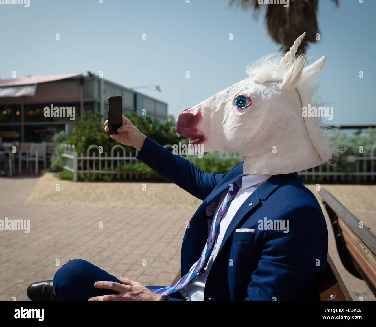 Young unusual man in comical mask and elegant suit makes a photo by phone. Funny unicorn is enjoying warm summer day. Strange guy sits on bench Stock Photo