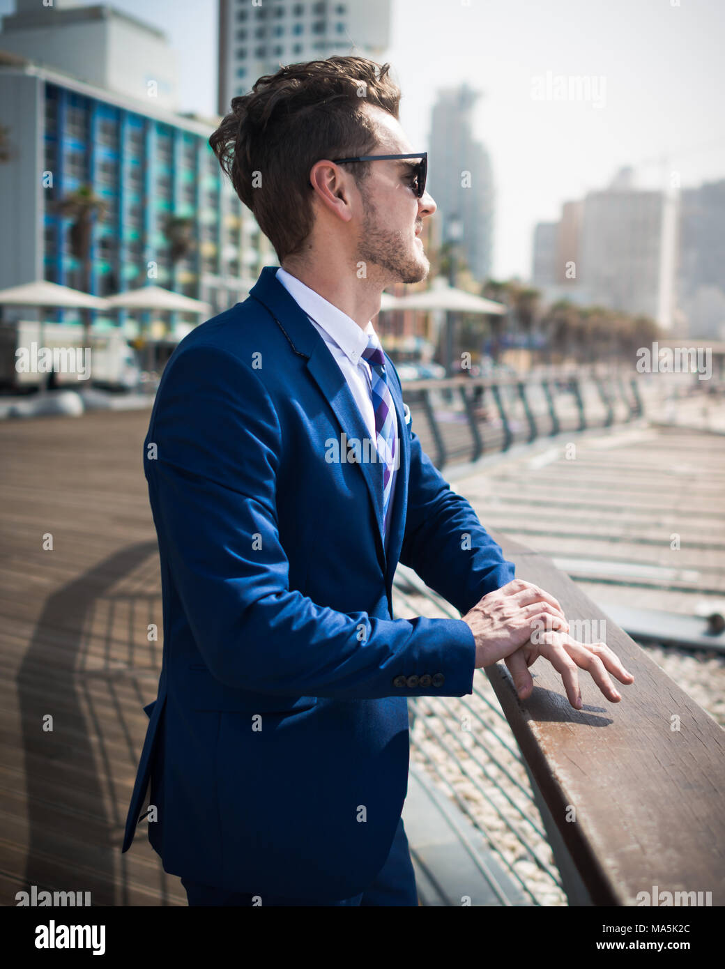Young man in suit and sunglass relaxing outdoors in warm sunny day. Successful manager on background of cityscape. Trendy guy stands on promenade Stock Photo
