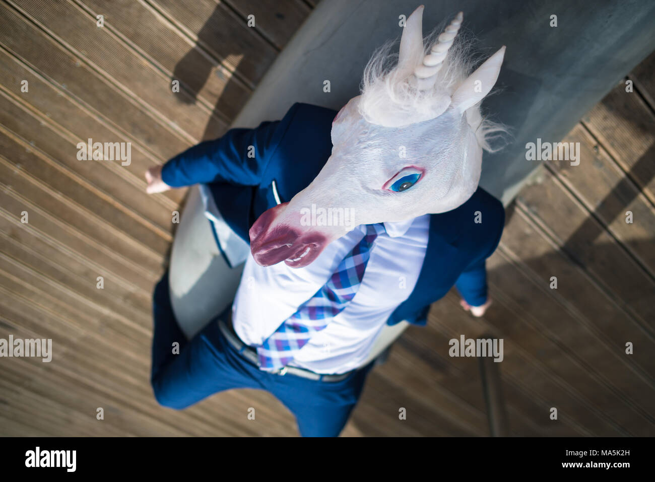 Young man in elegant suit lies under umbrellas on city waterfront. Unusual businessman in funny mask relaxes on vacation. Unicorn enjoying holidays Stock Photo