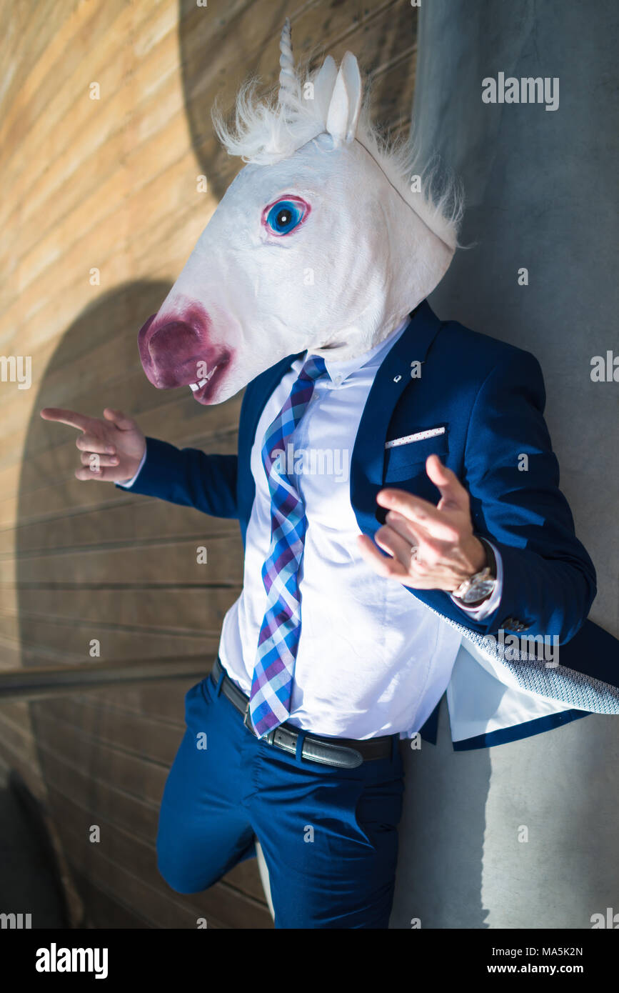 Young successful manager in elegant suit and funny mask active gesticulate. Unusual guy points to you. Unicorn spreads hands and relaxing on bench Stock Photo