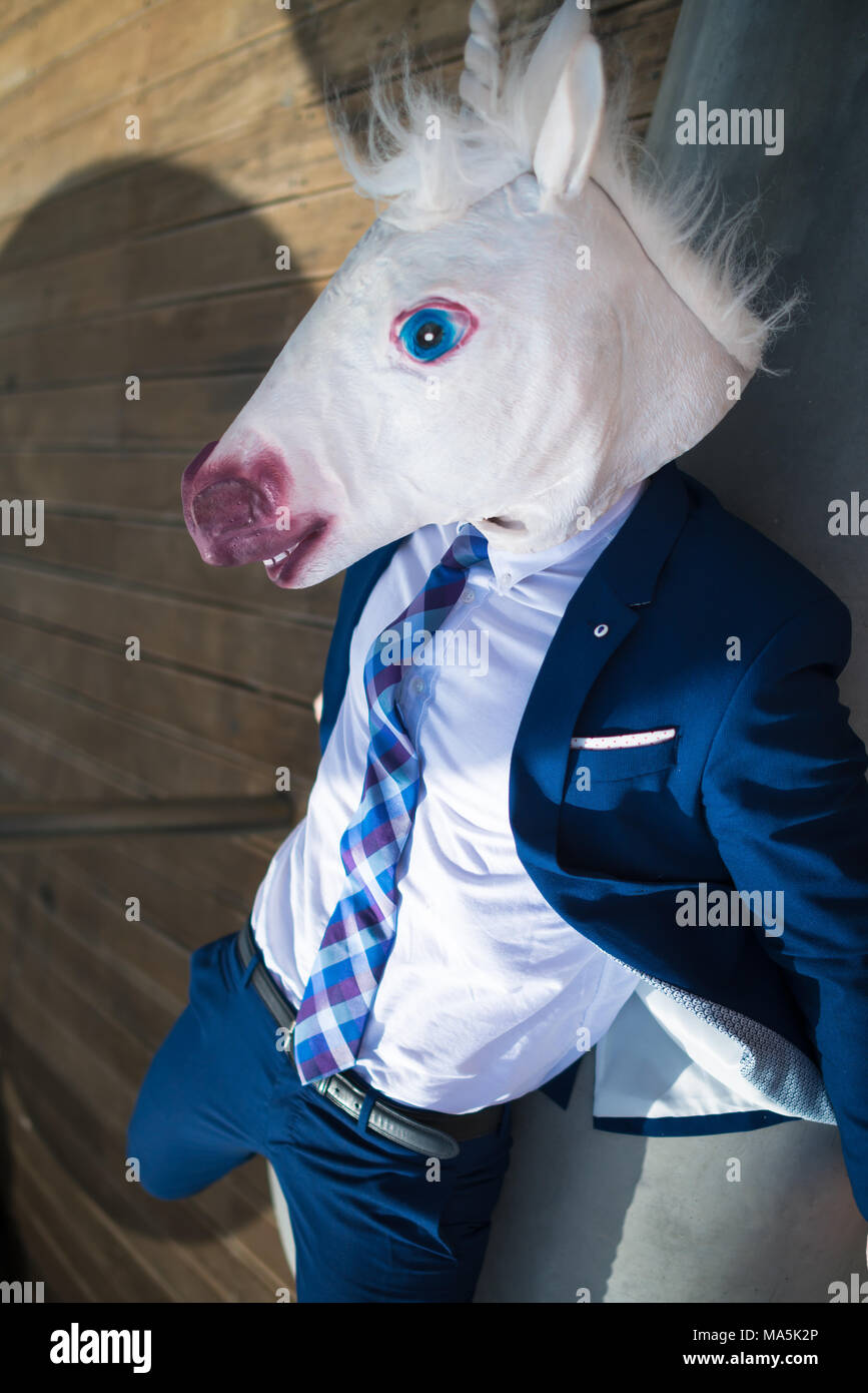 Very tired young manager in elegant suit and funny mask relaxes on bench. Unusual man has rest in warm summer day. Comical unicorn lies down Stock Photo