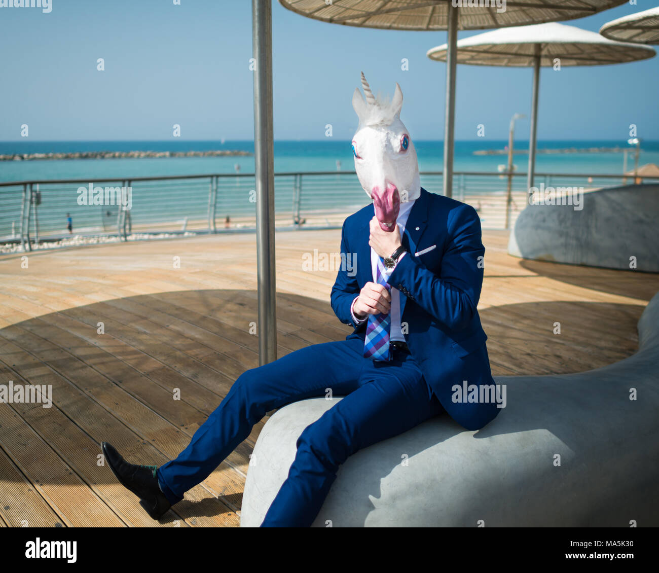 Young man in suit under stylish umbrellas on the city waterfront. Unusual traveler in comical mask. Funny unicorn sits at city promenade near sea Stock Photo