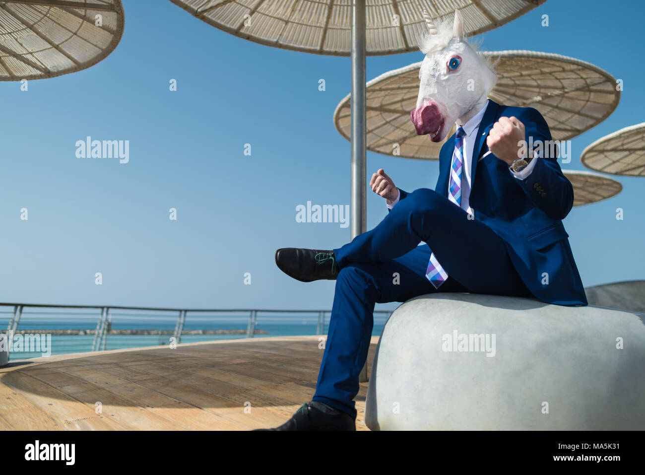 Young man in suit under umbrellas on city waterfront clenches fists. Unusual businessman in funny mask shows strength on promenade. Elegant unicorn Stock Photo