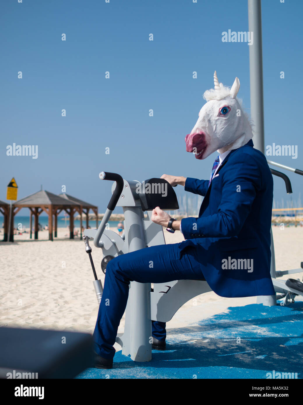 Funny athlete in suit and mask does exercises at sport ground near beachfront. Unusual man pumps the muscles. Unicorn is engaged in fitness outdoors Stock Photo