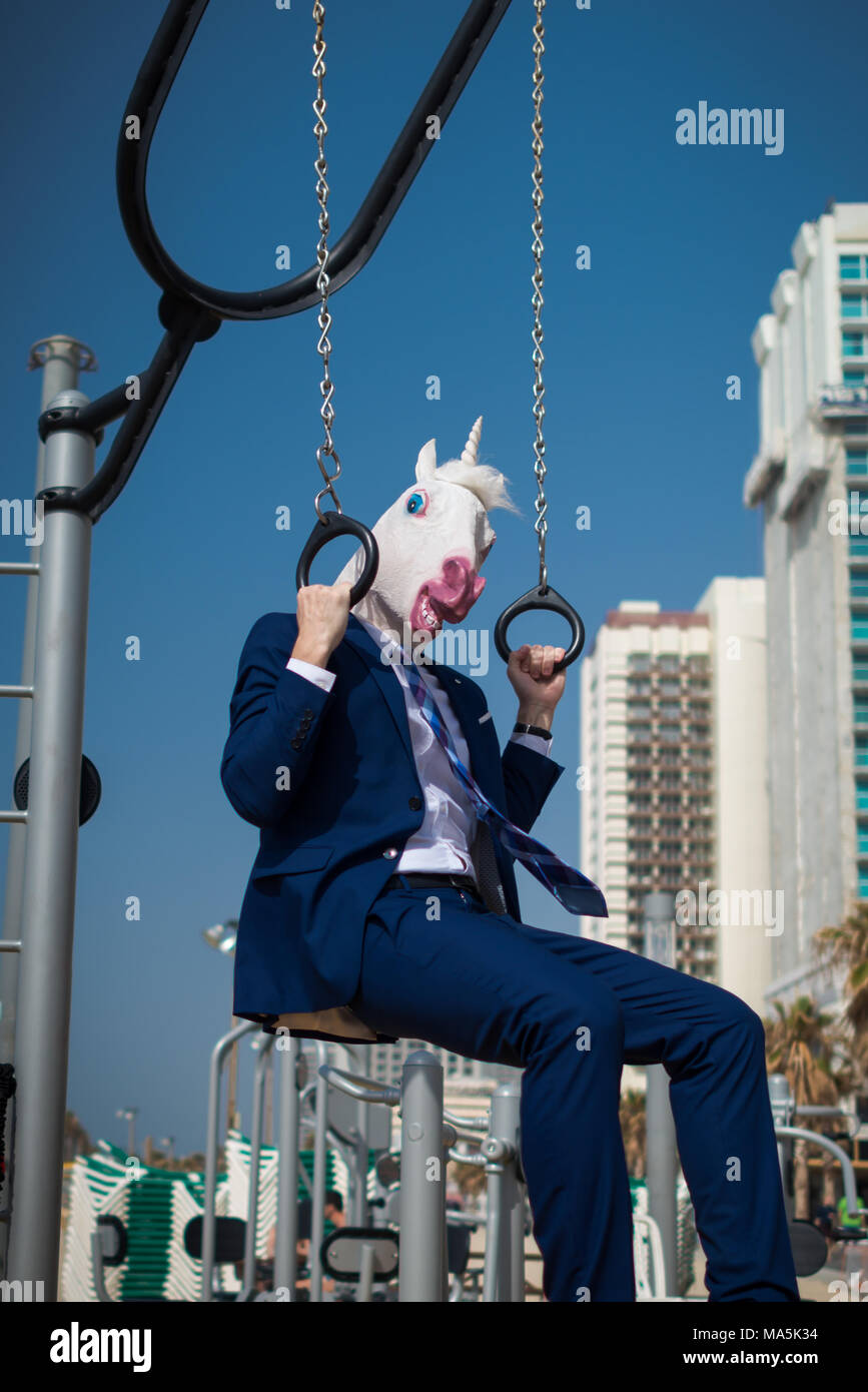 Funny fitness trainer in suit and mask does exercises at sport ground near beachfront. Unusual man pumps muscles. Unicorn engaged in fitness outdoors Stock Photo