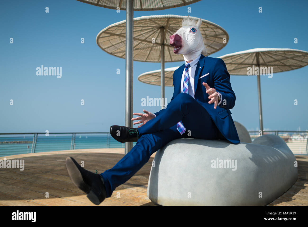 Young manager in suit under umbrellas spreads his hands. Unusual manager in comical mask is surprised. Funny unicorn sits at city promenade Stock Photo