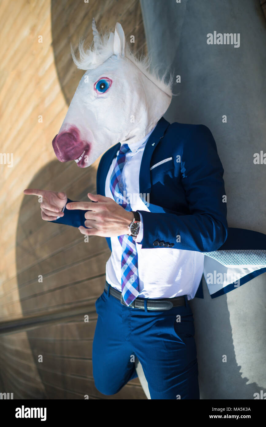Young cool man in suit and funny mask active gesticulate. Unusual manager points to you. Unicorn spreads hands at promenade and has relax on the bench Stock Photo