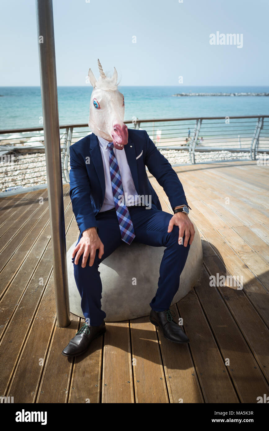 Strange guy in elegant suit sits on the city waterfront. Young unusual man in funny mask is resting on city promenade. Unicorn on background of sea Stock Photo
