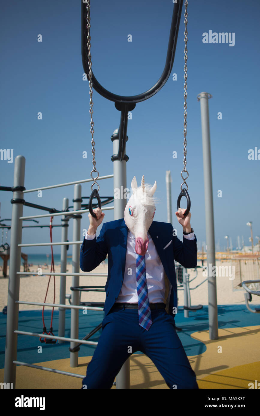 Funny businessman in suit and mask does exercises at sport ground near beach. Unusual strange man pumps muscles. Serious unicorn is engaged in fitness Stock Photo