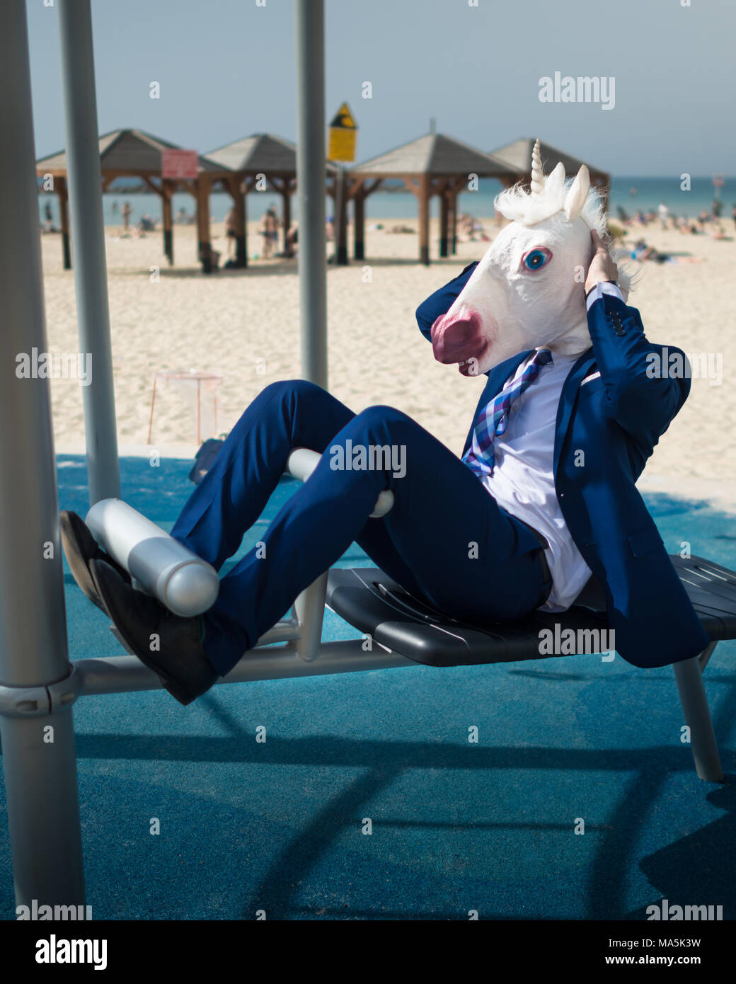 Comical man in suit and funny mask doing sports near beachfront. Unusual concentrated guy pumps muscles. Elegant unicorn fitness Stock Photo