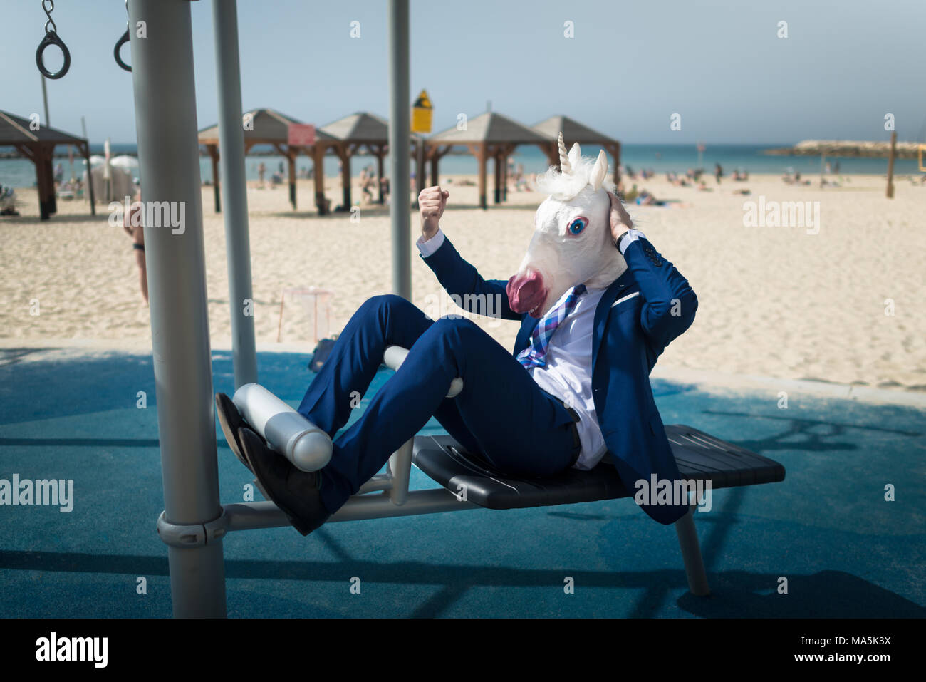 Unusual man in unicorn mask doing sport near beach. Freaky guy in suit pumps muscles and shows strength. Funny businessman fitness outdoors Stock Photo