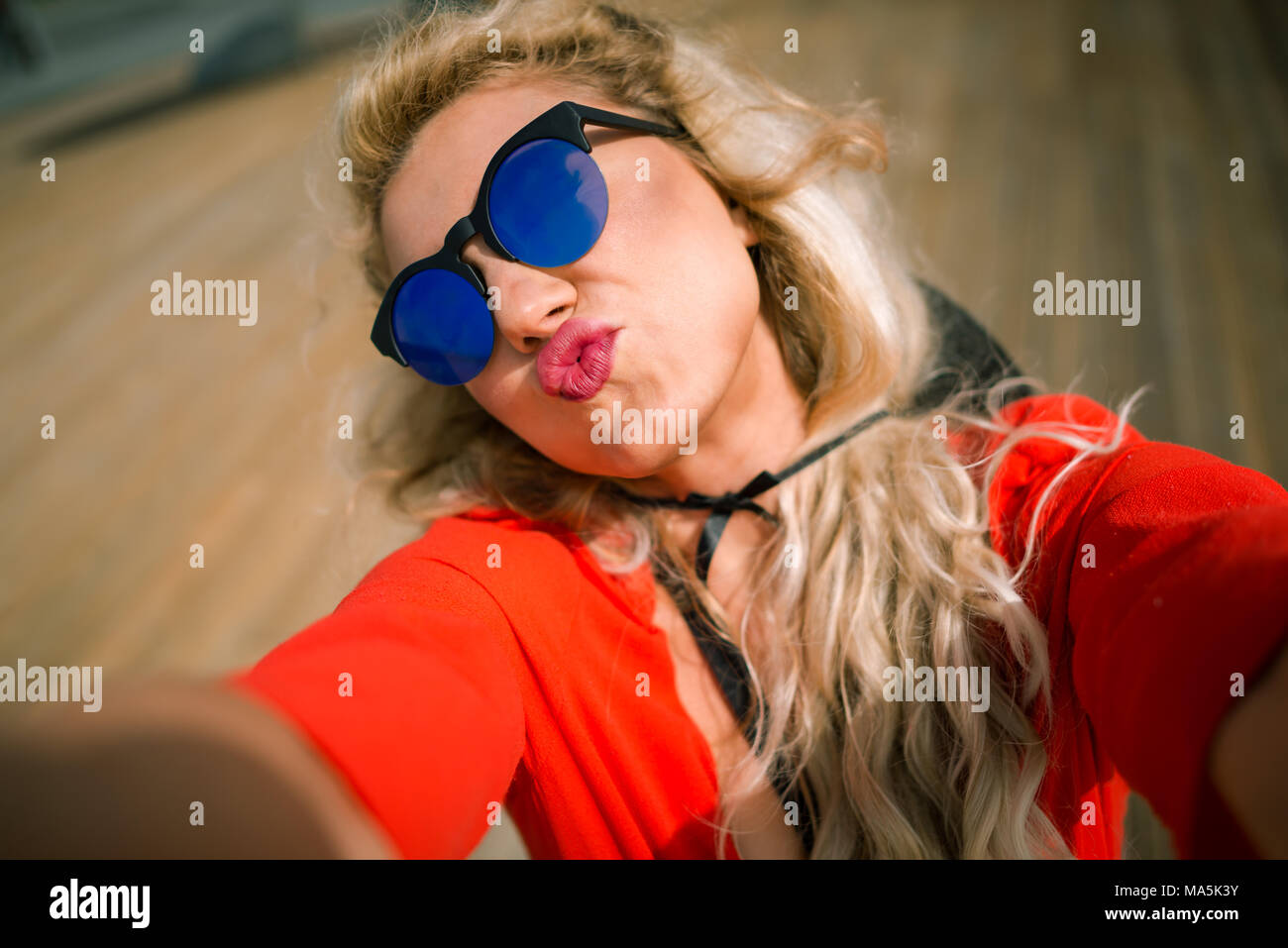 Close-up view of young beautiful woman in bright red dress shows a kiss at city waterfront. Elegant blonde girl with pretty face on wooden promenade Stock Photo