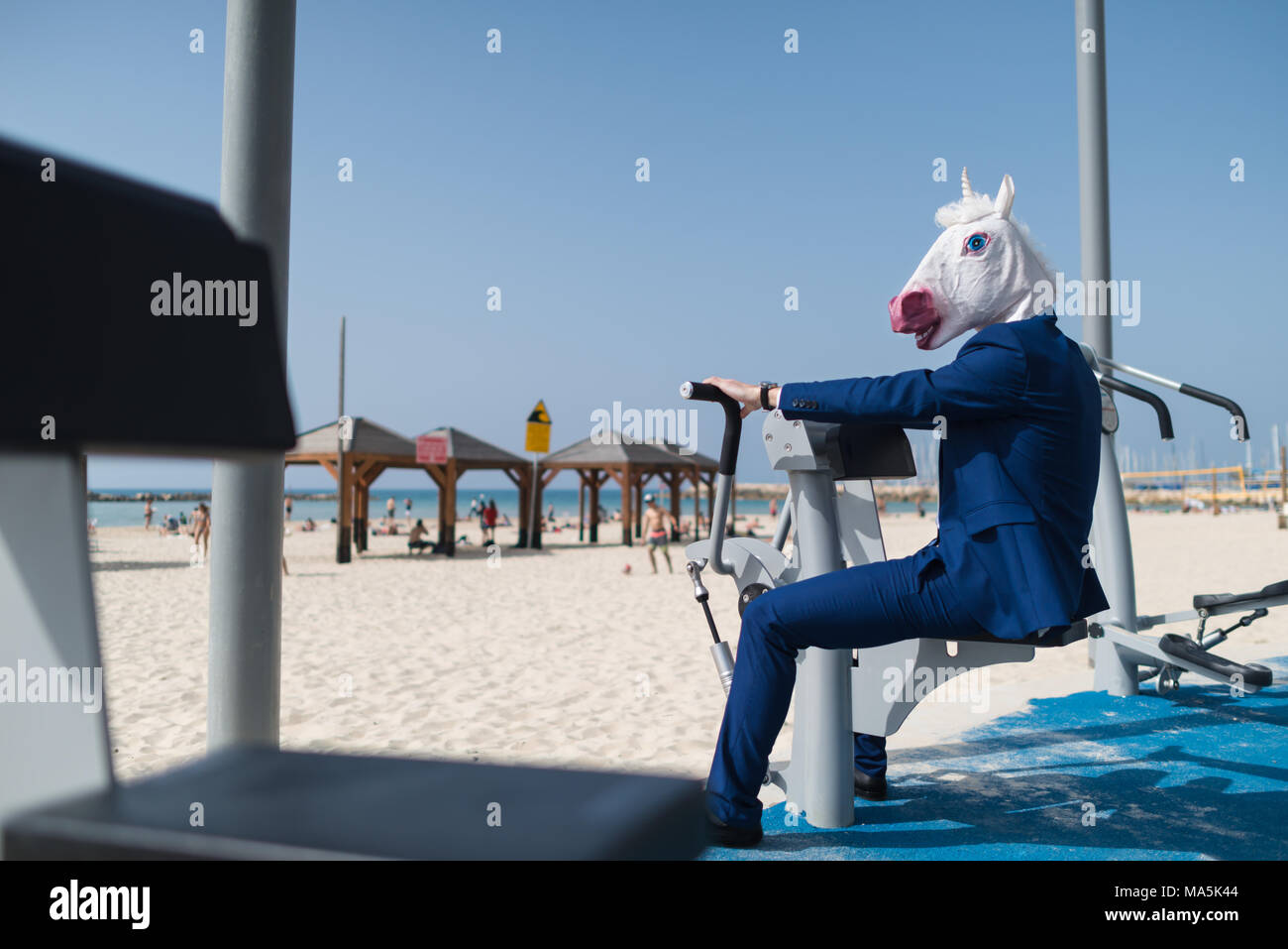 Funny sportsman in suit and mask does exercises at sport ground near beachfront. Unusual man pumps the muscles. Unicorn is engaged in fitness outdoors Stock Photo