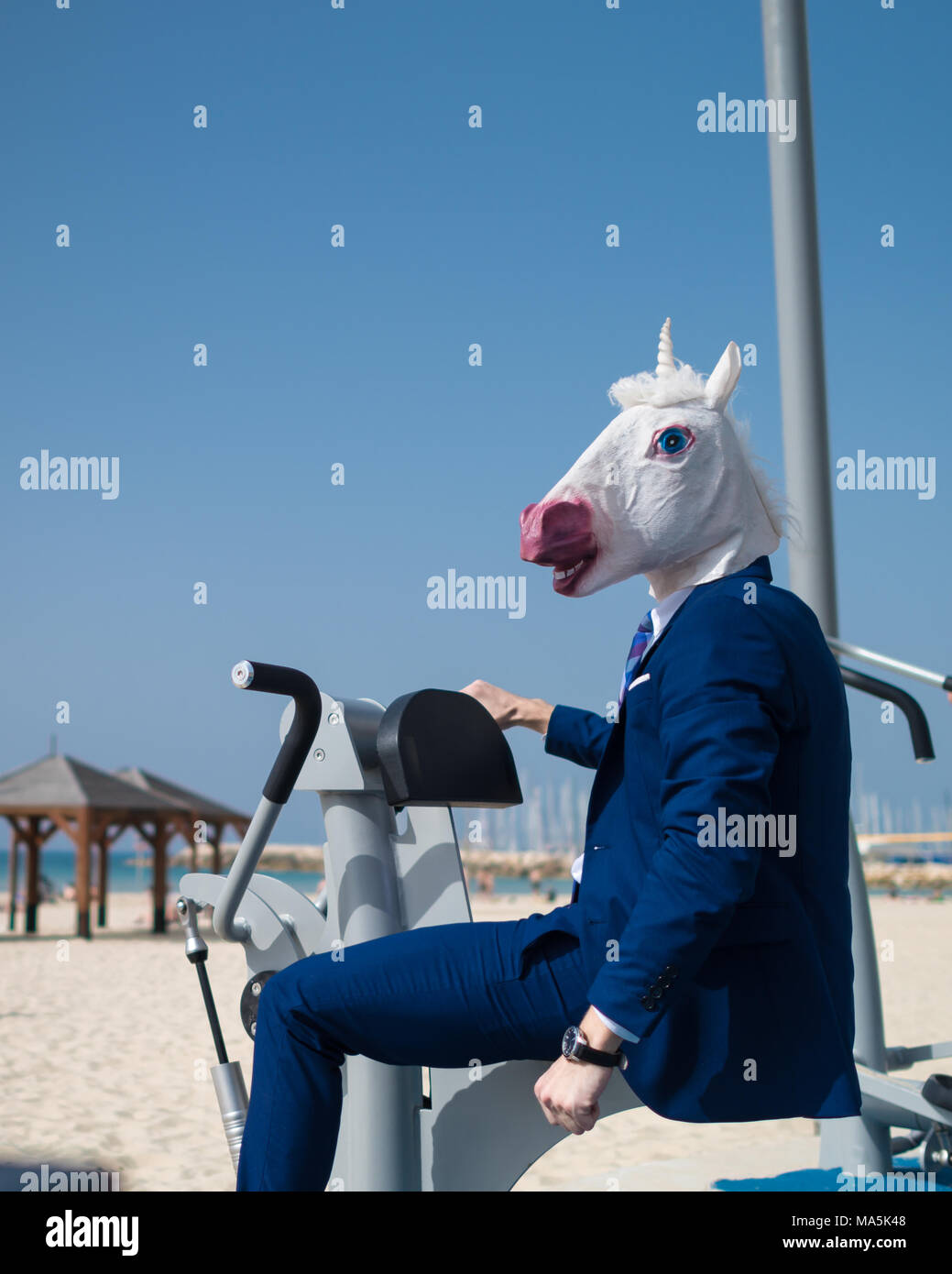 Strange guy in suit and mask does exercises at sport ground near beachfront. Unusual man pumps muscles. Funny unicorn is engaged in fitness outdoors Stock Photo