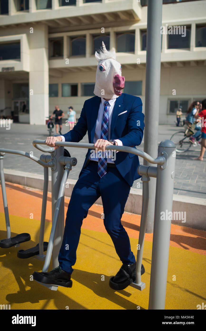 Freaky man in elegant suit and funny mask does sport exercises in sport ground near beachfront. Elegant unicorn is engaged in fitness outdoors Stock Photo