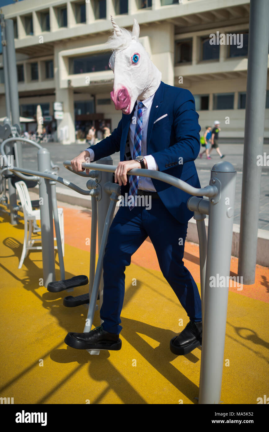 Funny man in suit and mask does sport exercises in sport ground near promenade. Unusual sportsman pumps muscles. Elegant unicorn is engaged in fitness Stock Photo