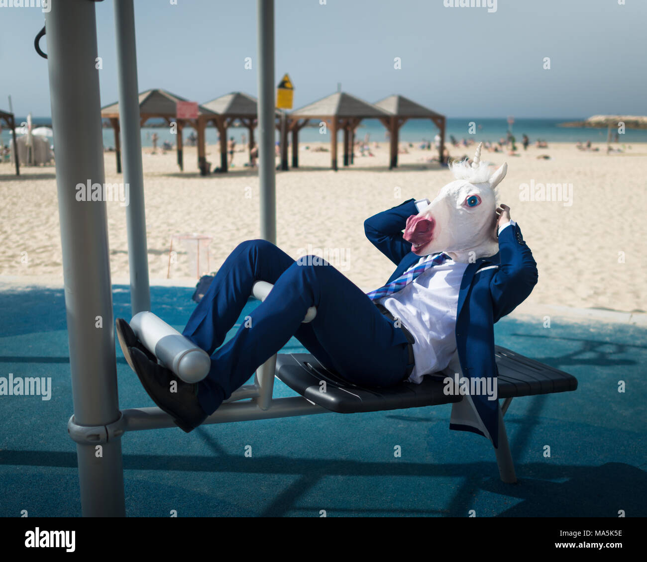 Freaky man in suit and mask is doing sport near beachfront. Unusual guy pumps press muscles. Elegant unicorn is engaged in fitness at the beach Stock Photo