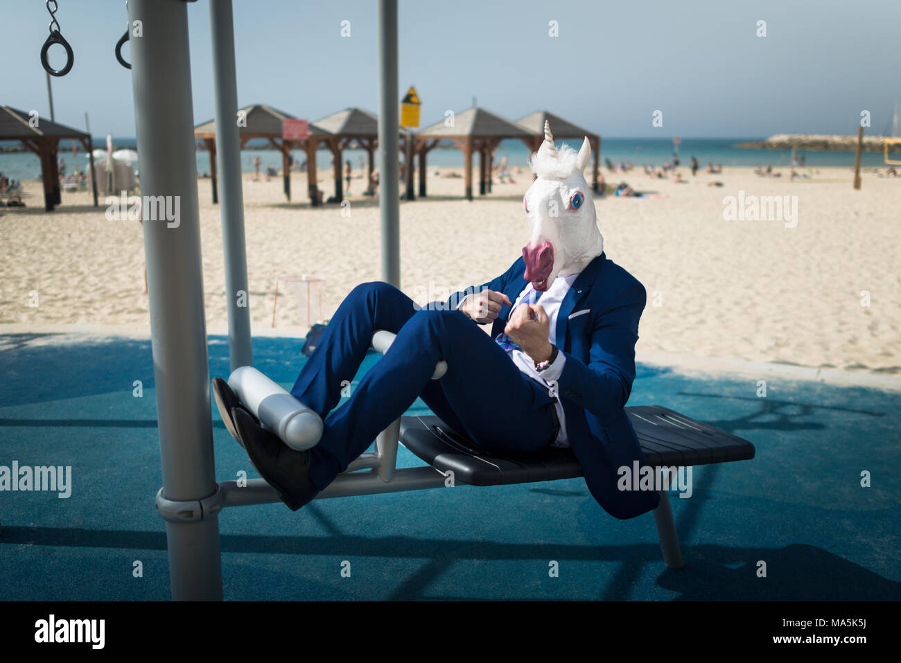Unusual man in suit and horse mask doing sport near beach. Freaky guy pumps muscles and shows strength. Funny unicorn fitness outdoors Stock Photo