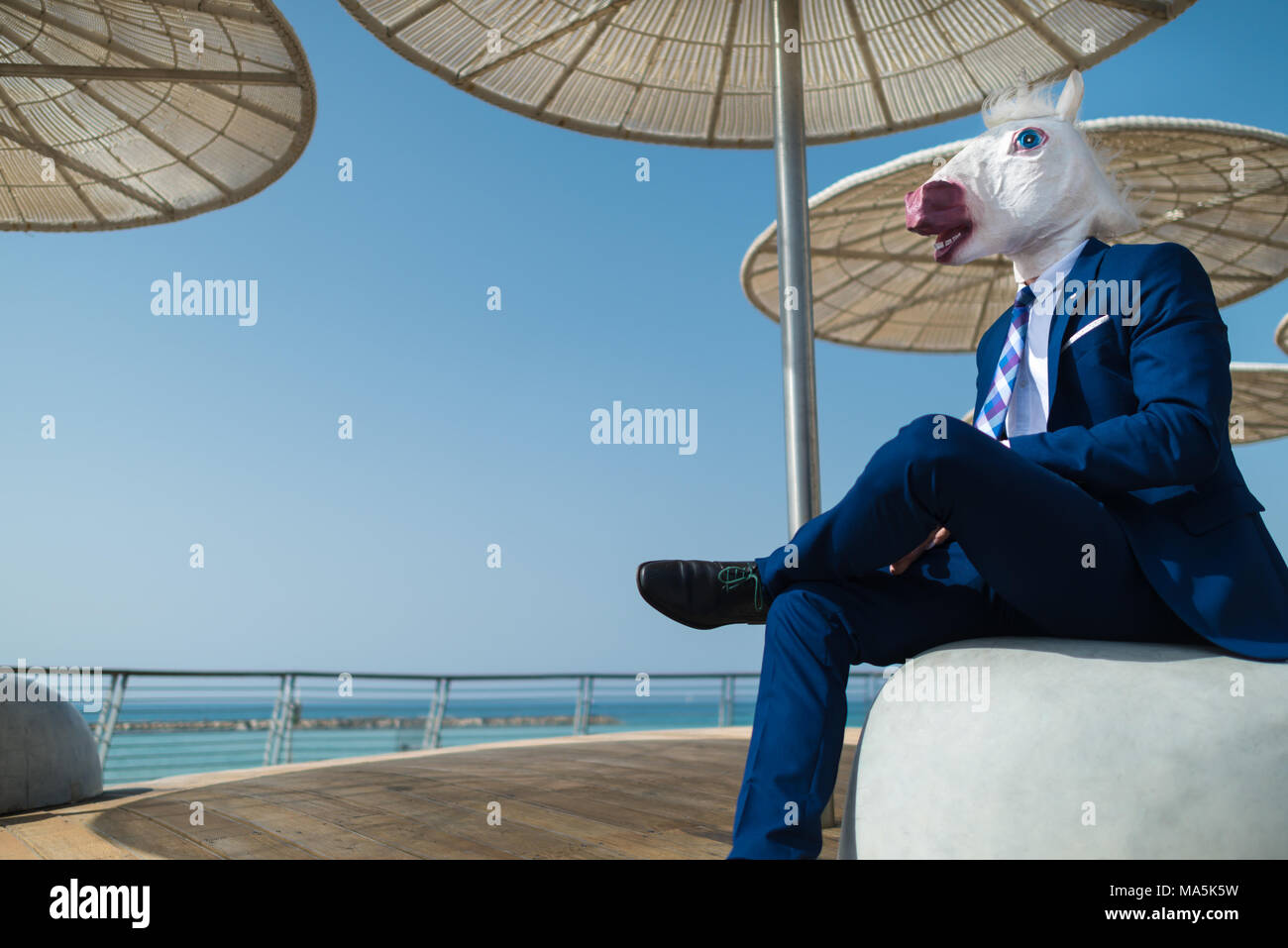 Young man in suit sits under stylish umbrellas on waterfront. Unusual businessman in funny mask relaxes on promenade. Unicorn is enjoying vacation Stock Photo