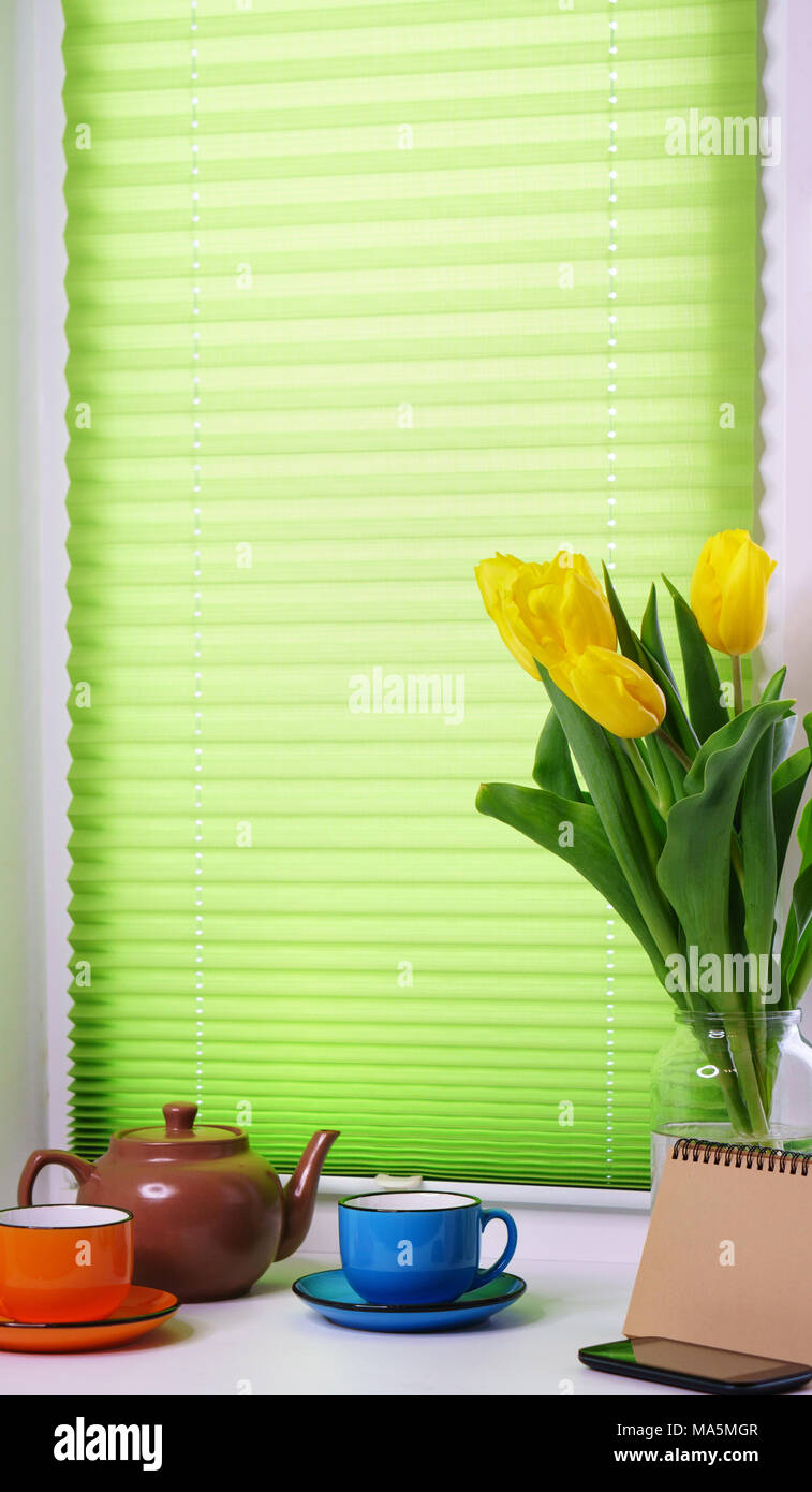 On windowsill there are yellow tulips, multi-colored tea cups, teapot, empty notebook for notes and cell phone. Stock Photo