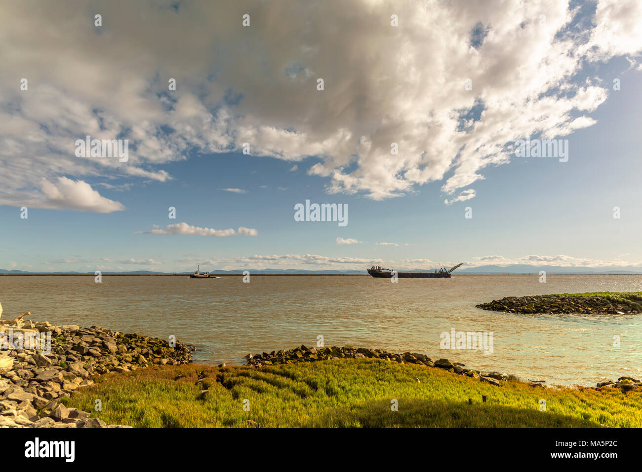 a view from the bank covered with grass and stones on the river with a boat towing a barge over the water, mountains in the background and a huge sky  Stock Photo