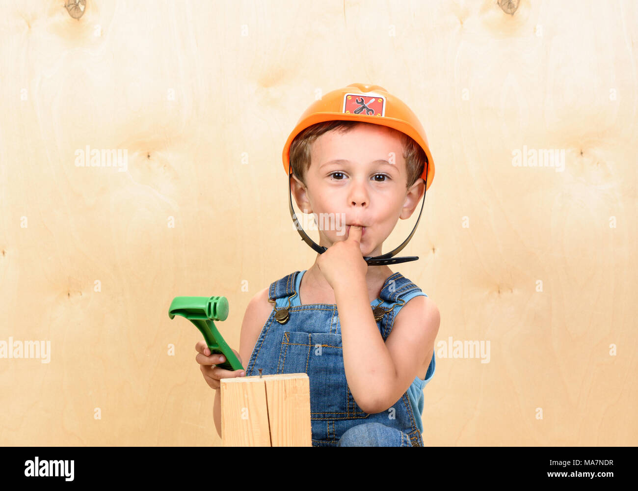 Kid boy as funny builder holding finger in mouth after hitting it with hammer Stock Photo