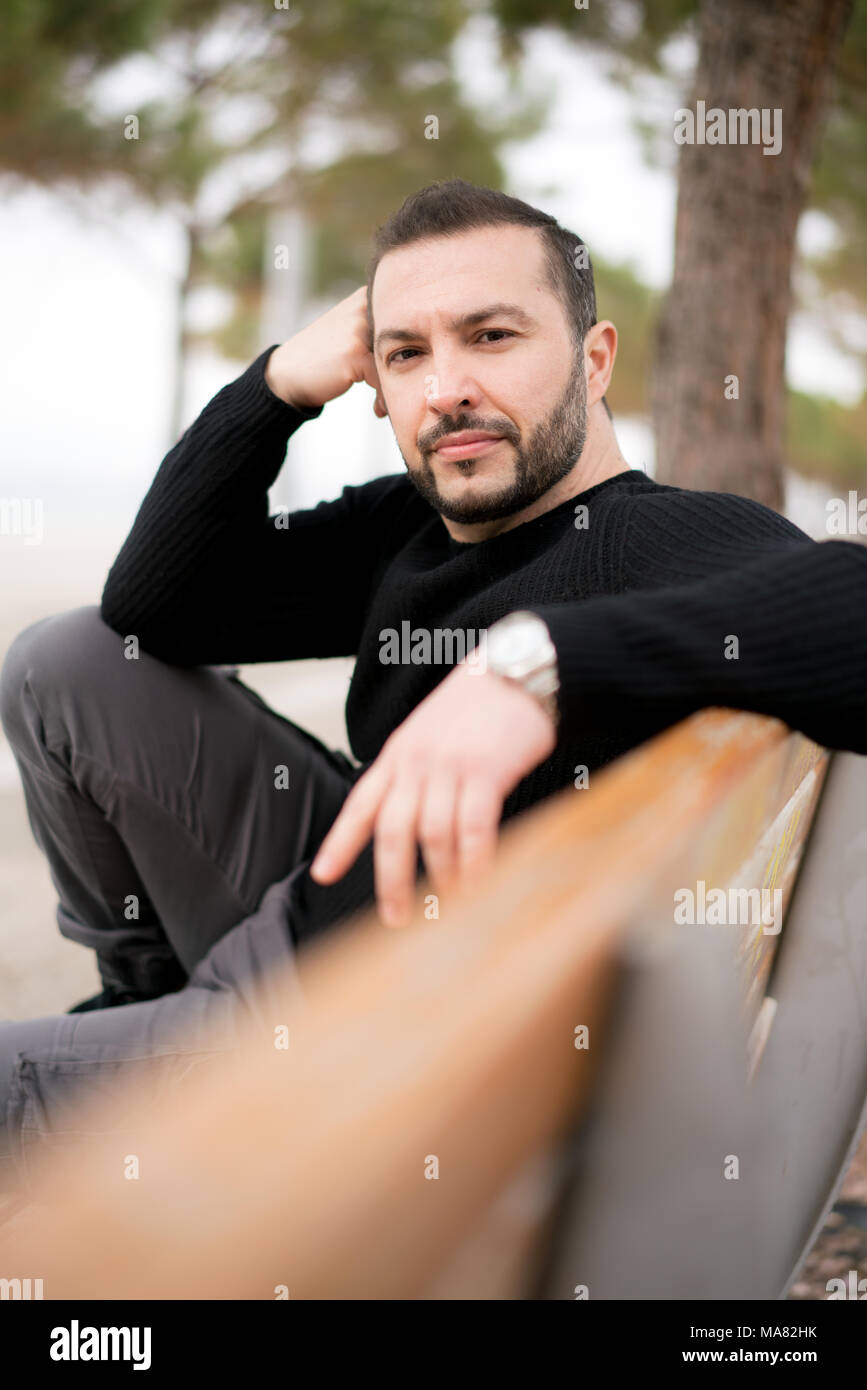 Portrait of Self Confident Man wearing casual clothes sitting on a bench at a park outdoors Stock Photo