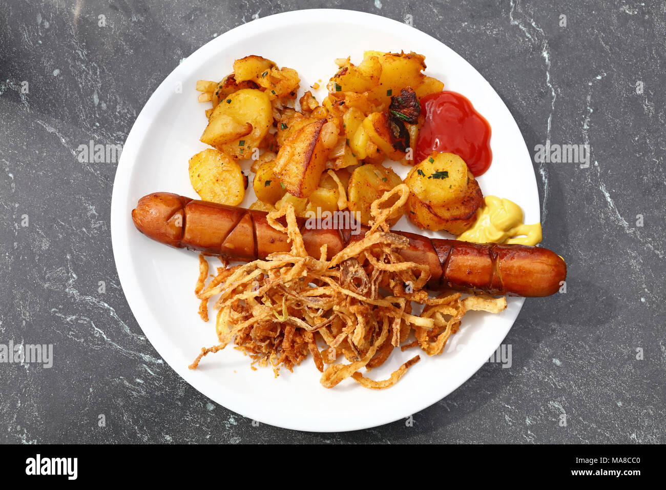 Close up portion of one big grilled sausage with homemade roasted potato, fried onion rings, ketchup and mustard on white plate over grey table, eleva Stock Photo