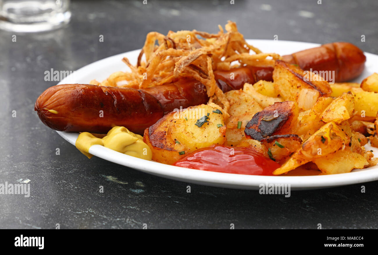 Close up portion of one big grilled sausage with homemade roasted potato, fried onion rings, ketchup and mustard on white plate over grey table, low a Stock Photo