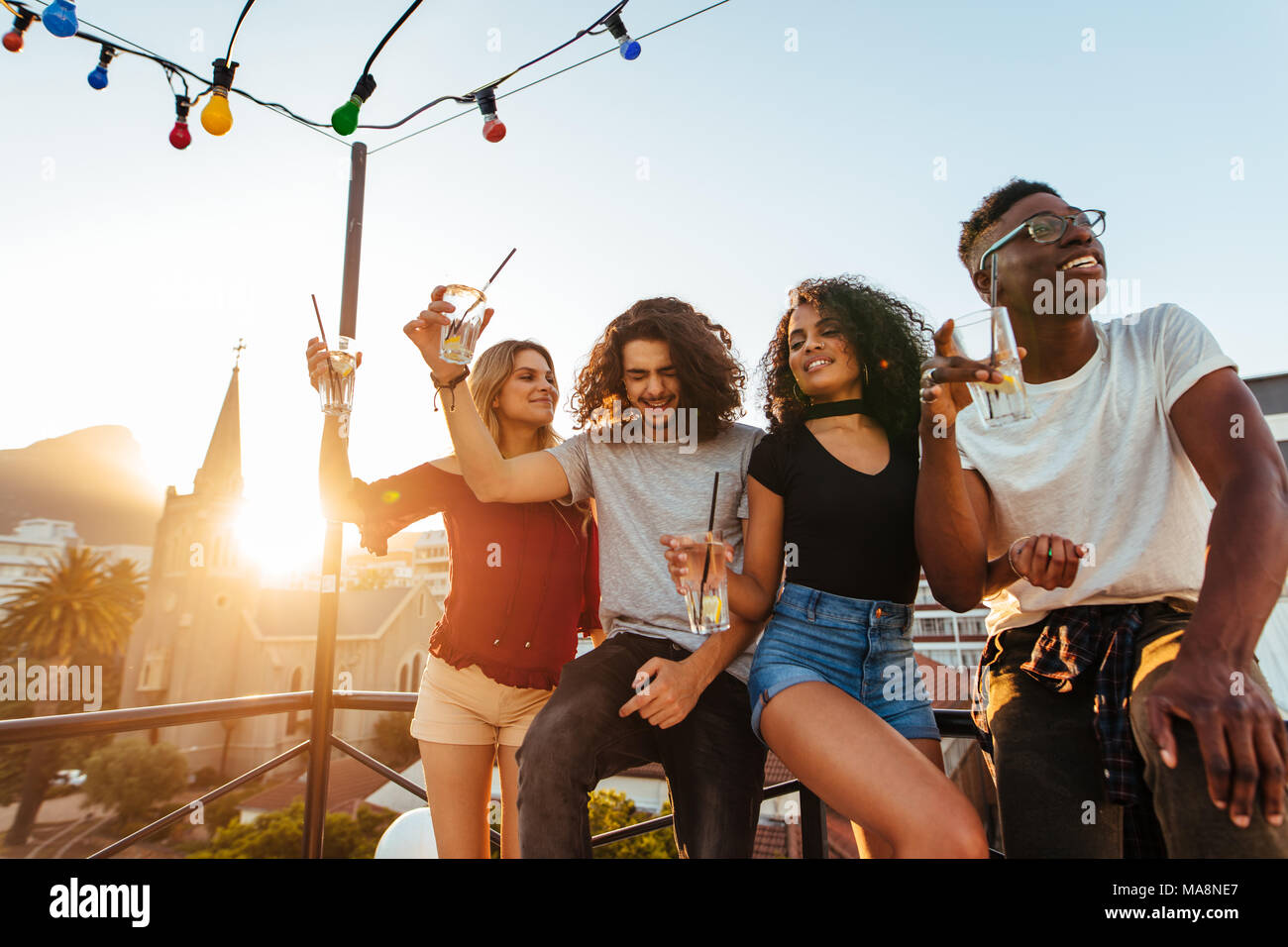 Group of young friends having fun, drinking and enjoying a evening on rooftop. Multiracial men and woman hanging out at rooftop party in evening. Stock Photo